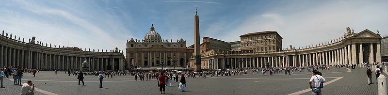 Panorama showing the facade of St Peter's at the centre with the arms of Berninis colonnade sweeping out on either side. It is midday and tourists are walking and taking photographs.
